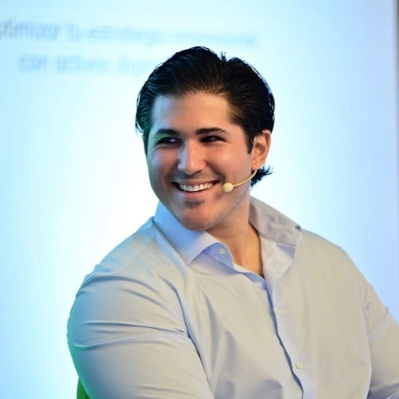 Smiling man with dark hair wearing a light blue shirt and headset microphone, speaking with a blurred blue background.